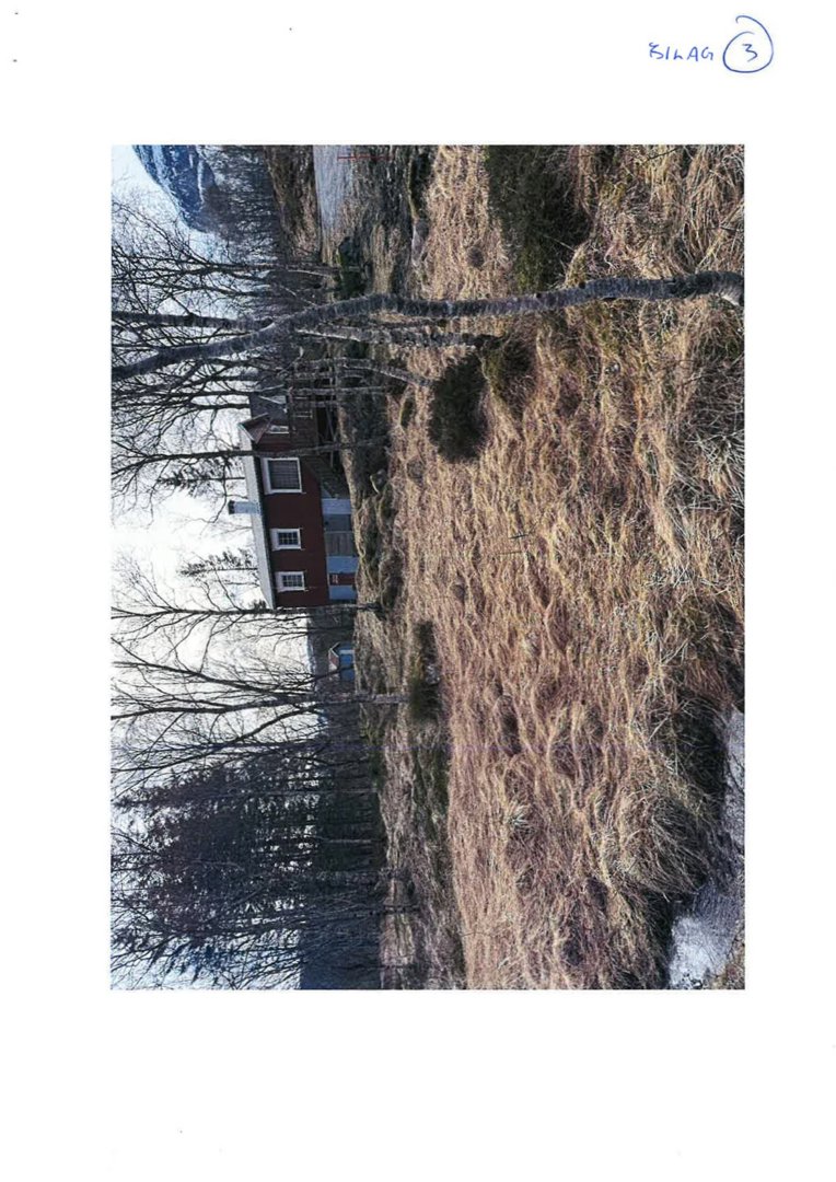Fotografi av en rød hytte eller hus sett fra siden, med skog og terreng i bakgrunnen.