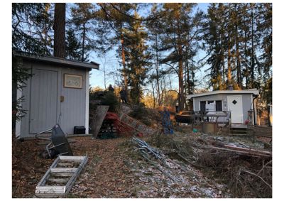 Fotografi av to småbygninger (sjekkestuer) på en tomt i skog, med utstyr og trær i forgrunnen.
