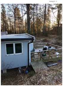 Fotografi av en liten hytte eller uthus sett fra siden, med et terrassedekke og omgivelser i skogen.