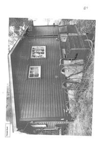 Fotografi av en mørk, trepanelert bygning sett fra siden (Fasade Nord). Bildet viser vinduer, takrenne, og omgivelser med trær og hage.