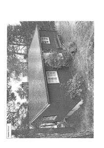 Sort-hvitt fotografi av en liten, mørk bygning (kanskje en hytte eller uthus) sett fra siden, omgitt av trær og busker.