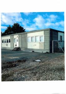 Fotografi av en lang, modulær bygning (kanskje container eller prefabrikkert hus) sett fra siden. Bygningen har flere vinduer, en dør med trapper, og er merket med tallene 4, 6 og 10. Det er en fotografering av en eksisterende bygning.