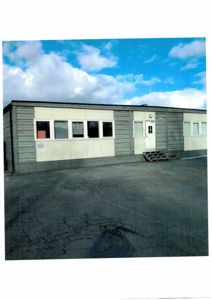 Fotografi av en grå, langbygget bygning (sannsynligvis en container eller modulbygg) sett fra siden med et asfaltert område foran.