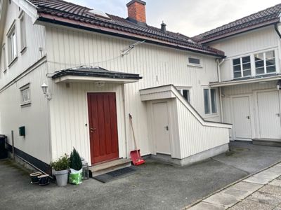 Fotografi av en hvit, trepanelert enebolig sett fra foran. Bildet viser fasaden med hoveddør, vinduer, tak, en liten utvendig trapp og en kjellerdør.