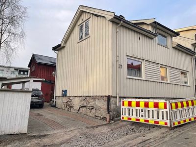 Fotografi av en beige farget, toetasjes bygning med hvite lister, sett fra siden. Bygningen har en steinfundament, vinduer på begge etasjer og en gavl med et vindu. Foran bygningen er det en gul og rød gjerde, og bakbygningen er synlig.