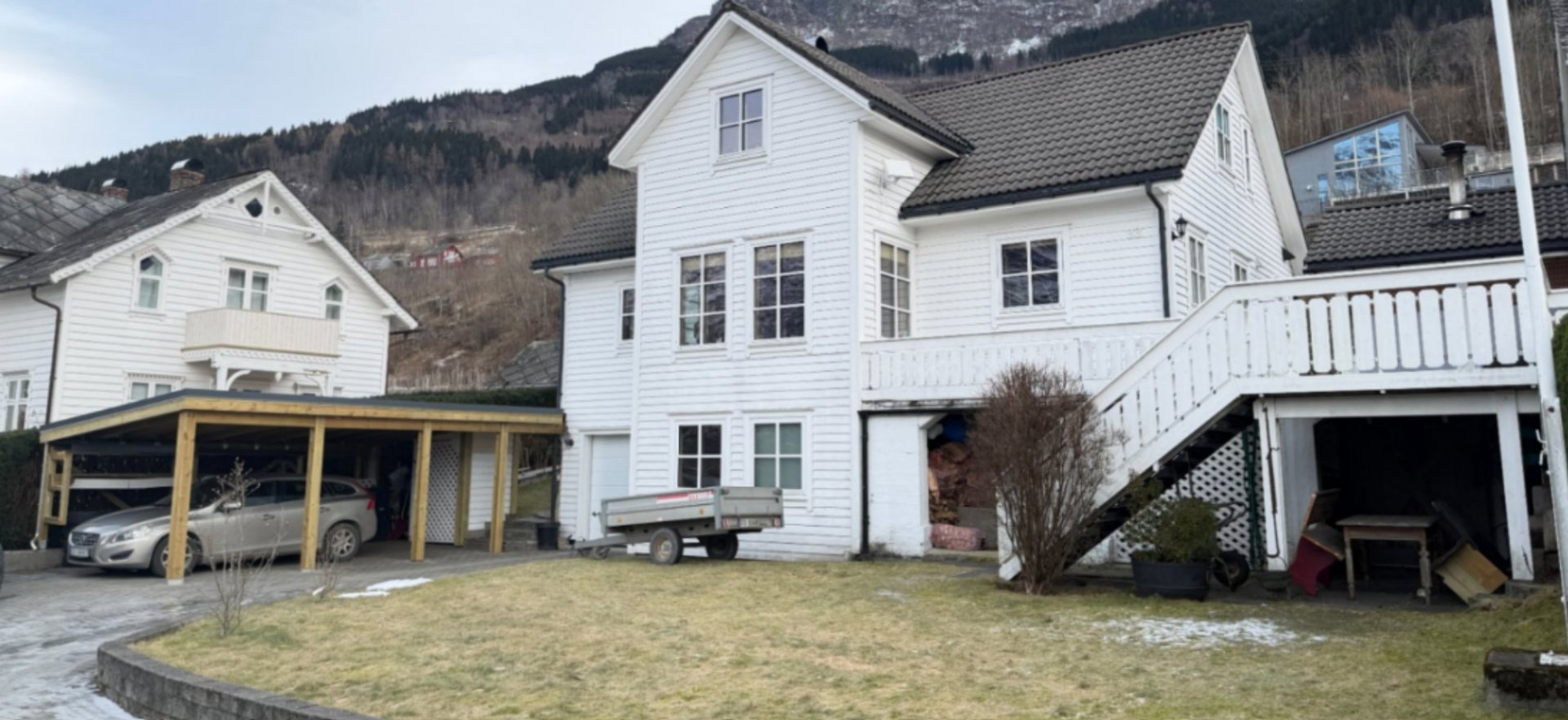 Fotografi av en hvit, trehuset bygning med en tilhørende carport og en trapp opp til en veranda. Bakgrunnen viser en fjellside.