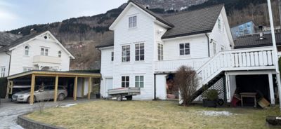 Fotografi av en hvit, trehuset bygning med en tilhørende carport og en trapp opp til en veranda. Bakgrunnen viser en fjellside.