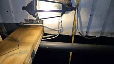 Fotografi av en utvendig lysarmatur montert på en hvit fasade, med en gul målebånd som henger ned langs veggen.