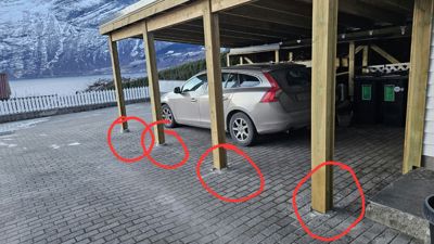Fotografi av en ferdigbygd carport med en bil parkert under. Bildet viser også en del av omgivelsene, inkludert vann og fjell i bakgrunnen. Det er røde sirkler tegnet over fundamentene under søyler.