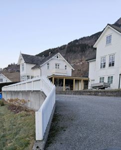 Fotografi av en hvit, to- eller treetasjes bygning (hus) sett fra en vinkel. Bildet viser fasaden, en tilhørende garasje/carport, en hvit hekk, en grusvei og bakgrunnen med en skråning.