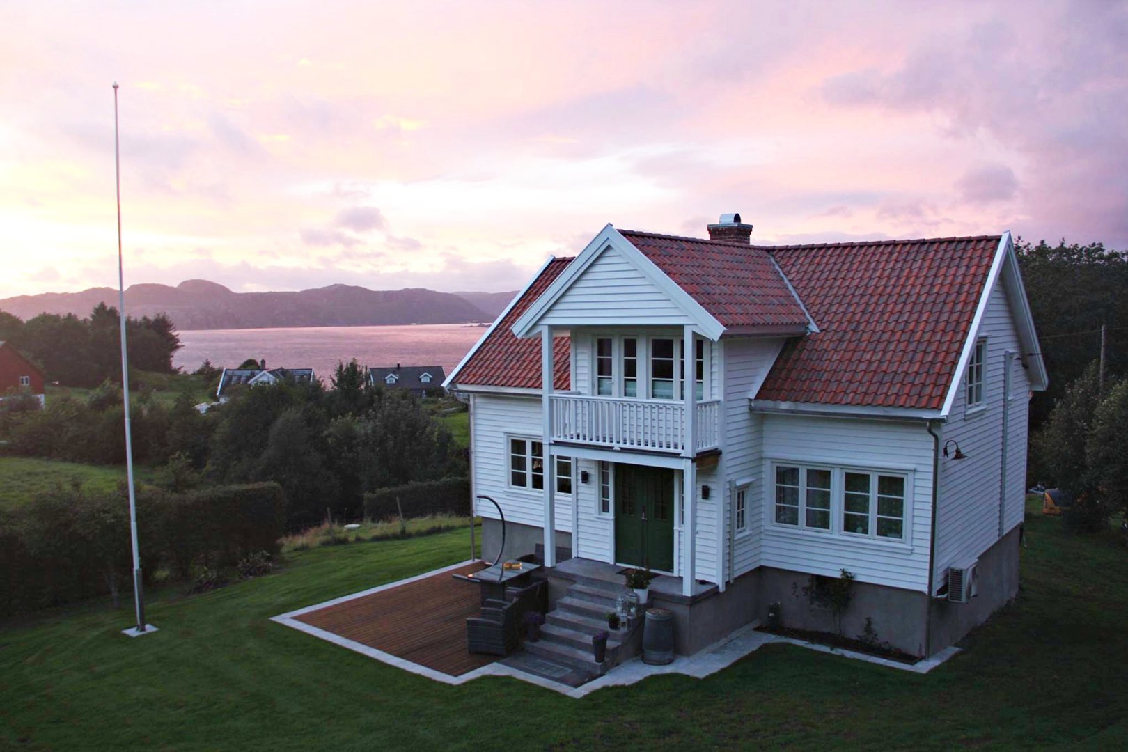 Fotografi av en hvit villa med rødt tak, sett fra en høy vinkel mot bakgrunn av fjord og fjell.