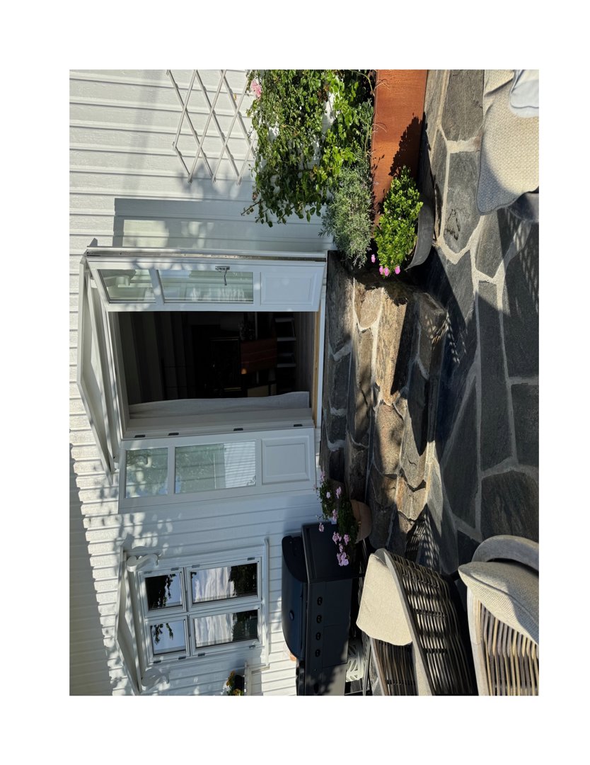 Fotografi av en hvit utvendig fasade med en åpen hvit dør og en vindu under. Bildet viser også en steinlagt terrasse med planter og utemøbler.