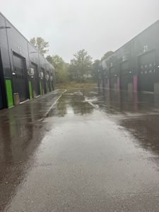 Fotografi av en rekke med garasjer eller lagerlokaler sett fra inngangsplassen. Bildet viser fasadene på bygningene, nummerering (f.eks. 15, 21, 22), dører, vinduer og den våte asfalten foran.