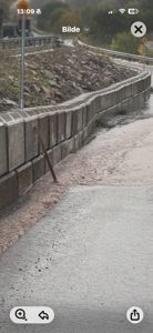 Fotografi av en betongmur langs en vei eller gangvei, med steinbakke og hekk i bakgrunnen.