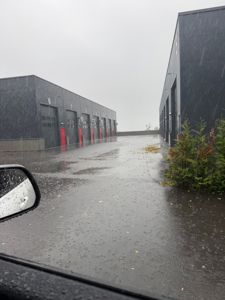 Fotografi av en industriell eller logistisk anlegg med flere store garasjeporter (merket 6, 7, 8 osv.) sett fra en bil under regn.