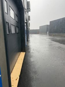Fotografi av en industriell eller lagerbygning sett fra utsiden under regn. Bildet viser fasaden, en betongplassering og en del av nabolaget.