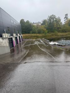 Fotografi av en industriell bygning med flere dører og luftbehandlingsanlegg, sett fra en fuktig asfaltert overflate mot en elv eller vannløp.