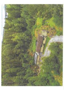 Luftfoto av en eiendom med to bygninger (hus og uthus) plassert på en skråning i skog.