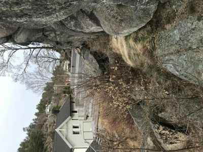 Fotografi av en hvit villa med mørk takflate, plassert i et kupert terreng med store granittblokker og trær.