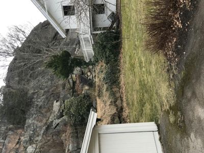 Fotografi av en hvit villa bygget inn i en steinvegg, sett fra bakgården eller siden. Bildet viser også en del av en garasje eller uthus i forgrunnen.