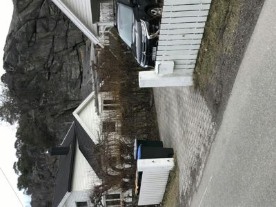 Fotografi av en eksisterende bygning (hus) sett fra siden, med en bil parkert foran og en steil fjellvegg i bakgrunnen.