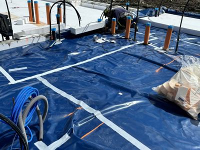 Fotografi av en byggeplass som viser arbeid med fundamentet, inkludert blå dampsperrmembran, isolasjonspiller og armering.