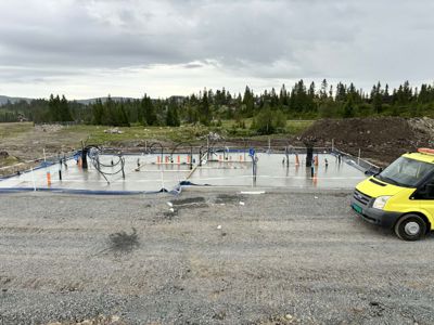 Fotografi av en byggeplass som viser en ferdigstilt betongfundament med utlagte rør og stålanker. I bakgrunnen er det skog og fjell.