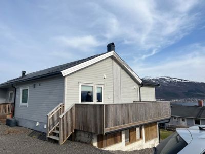 Fotografi av en eksisterende enebolig sett fra siden, med en stor terrasse, trapp og en bakgrunn med fjell og vann.