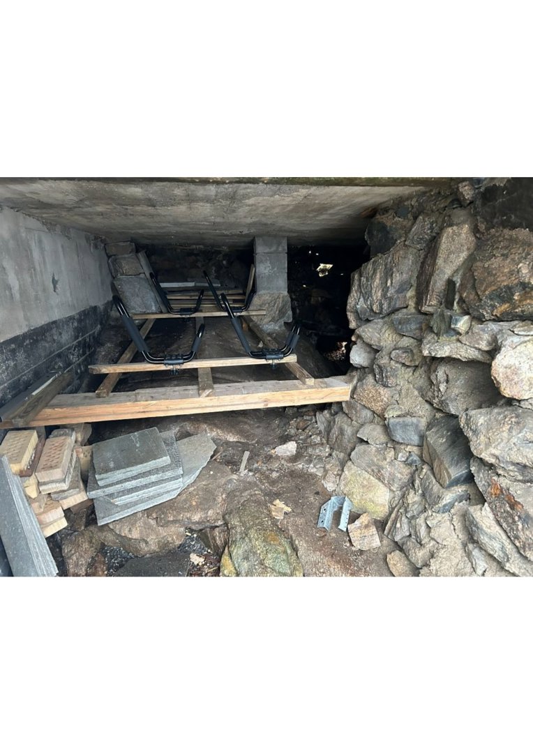 Fotografi av en underjordisk eller halvunderjordisk konstruksjon, sannsynligvis en kjeller eller graving, med synlig steinmur, betongtak og midlertidig tregulv.