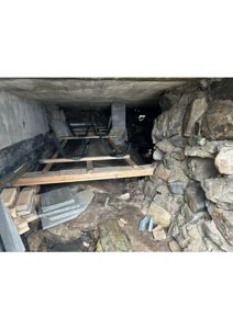 Fotografi av en underjordisk eller halvunderjordisk konstruksjon, sannsynligvis en kjeller eller graving, med synlig steinmur, betongtak og midlertidig tregulv.