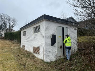 Fotografi av en hvitmalt, liten bygning (sannsynligvis en transformatorstasjon eller lignende) sett fra siden. En person i gul vest står ved døren.