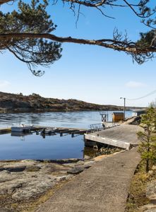 Fotografi av en kai, brygge og en liten båthavn ved en kyst med vann og kupert terreng i bakgrunnen.