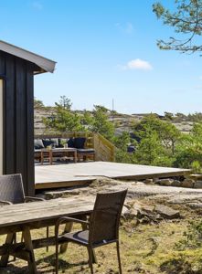 Fotografi av en ute terrasse med møbler foran et mørkt trehus, sett fra bakken mot utsikt over kystlandskap.