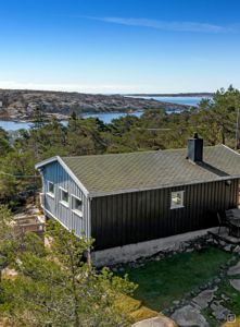 Fotografi av en hytte sett fra et høyere utsiktspunkt, med hav og kystlandskap i bakgrunnen.