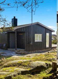 Fotografi av en mørk, trepanelt hytte eller uthus sett fra siden, med en steinete og mosete hage i forgrunnen.