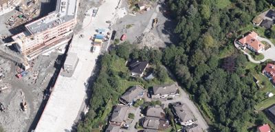 Luftfoto av et byggefelt med en delvis ferdig bygning, en stor grusflate, og omgivende bebyggelse og skog.