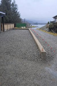 Fotografi av en uteareal med grus, en betongmur og en vannkant i bakgrunnen.