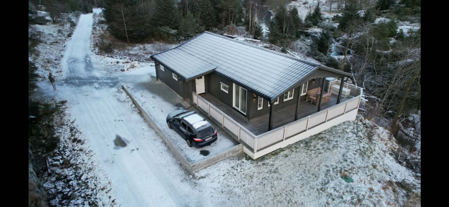 Fotografi av en ferdigbygd eller ferdigstilt enebolig sett fra luften (dronebilde). Bildet viser huset i sin helhet med omgivelsene, inkludert snødekke, vei og bil.