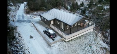 Fotografi av en ferdigbygd eller ferdigstilt enebolig sett fra luften (dronebilde). Bildet viser huset i sin helhet med omgivelsene, inkludert snødekke, vei og bil.