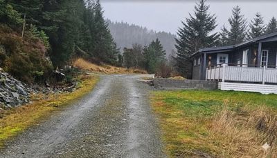 Fotografi av en grusvei som fører opp mot en hytte i et skogkledt landskap.