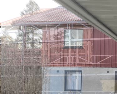 Fotografi av en bygning under oppføring eller renovering, sett gjennom et vindu. Bildet viser fasaden med rødt tak, vinduer og deler av konstruksjonen.