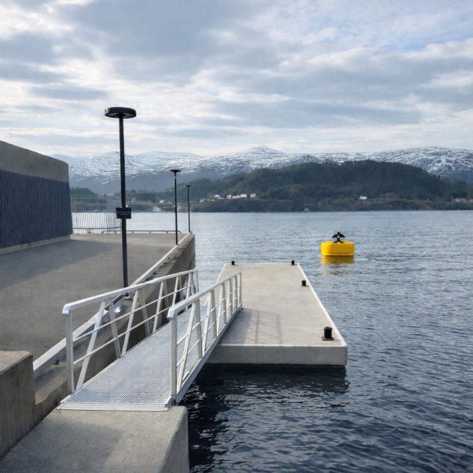 Fotografi av en nybygd kai med ramp og lysstolper ved vannet, med fjell i bakgrunnen.