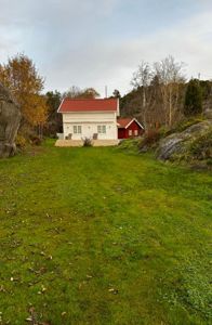 Fotografi av en hvit hytte med rødt tak og en mindre rød uthus, sett fra en grønn hage mot bakgrunn av skog og fjell.