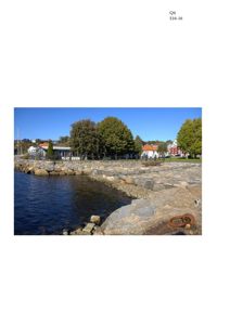 Fotografi av en kystlinje med steinmur, vann og hus i bakgrunnen.