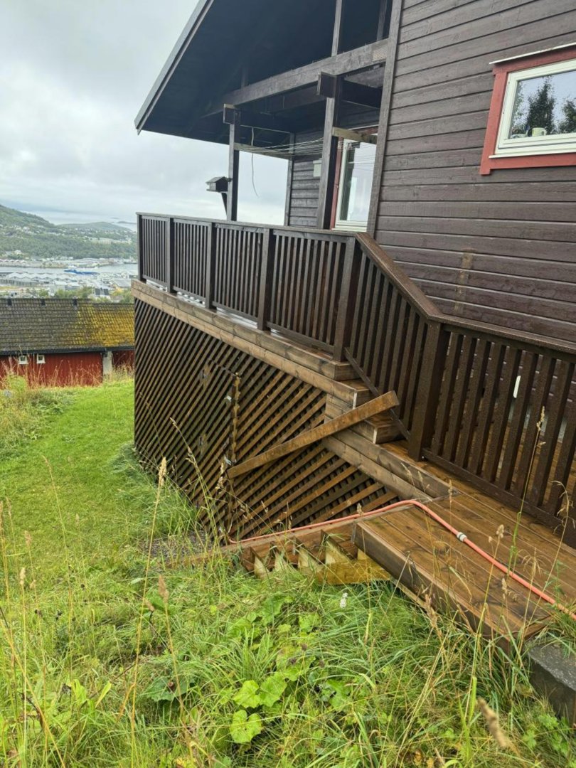 Fotografi av en mørk trehytte sett fra siden, med fokus på en trapp og en veranda som går nedover en skråning.
