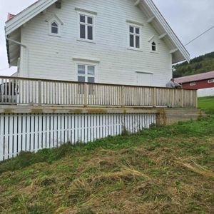 Fotografi av en hvit, toetasjes trehytte sett fra siden. Bildet viser fasaden, etasjenes vinduer, en utvendig terrasse med balustrade og en hvit panelvegg under terrassen. I bakgrunnen er det en rød bygning og en skogkledde bakke.