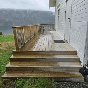 Fotografi av en ferdigbygd terrasse med trinn og hekk foran en hvit bygning, med utsikt over vann og fjell.