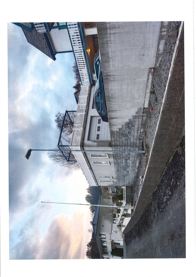 Fotografi av en eksisterende eller ferdig bygning sett fra siden, med bil og gatelys i forgrunnen.