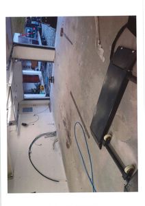 Fotografi av en garasje eller lagerhall med en heis, kabler på veggen og en dør som er åpen mot en hage.