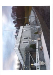 Fotografi av en eksisterende bygning sett fra siden, med omgivelser som trapp, hekk og trær.